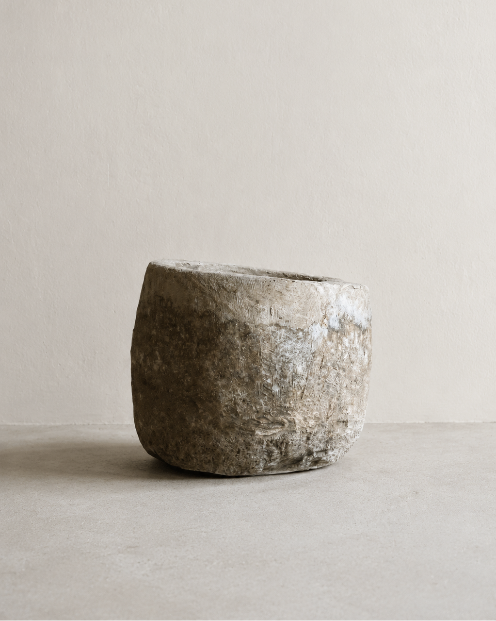 A rustic, hand-crafted stone planter pot with a rough, textured surface, displayed on a neutral concrete surface against a minimalist beige wall.